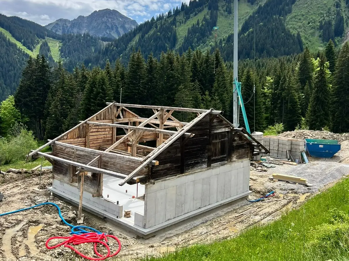 Vieux chalet en bois posé sur son nouveau soubassement en béton, grue du chantier à côté, montagnes au fond